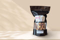 A tan background behind a black resealable bag. Inside the bag is chocolate and caramel coated kettle corn. 