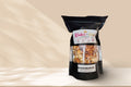 A black stand-up pouch of Kakesthe Great Karamel Machiatto popcorn, featuring a clear window that displays the golden-brown popcorn inside, set against a warm beige background with soft shadow overlays