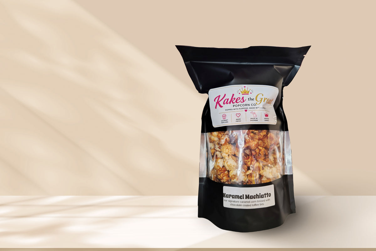 A black stand-up pouch of Kakesthe Great Karamel Machiatto popcorn, featuring a clear window that displays the golden-brown popcorn inside, set against a warm beige background with soft shadow overlays