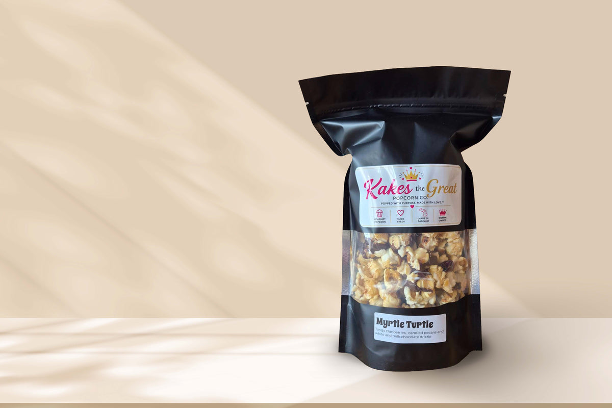 Cranberries, chocolate, and candied pecans can be seen mixed with Kakes the Great's kettle corn inside a black bag. The bag sits on a beige countertop and is in front of a tan background. 