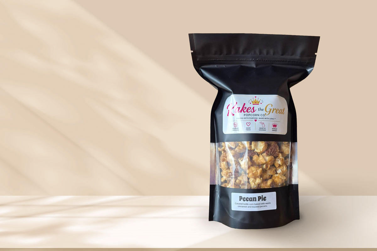 A black resealable stand-up pouch of Kakes of Great popcorn mix, flavor Pecan Pie, standing on a light beige surface against a matching beige background with soft shadow overlays.