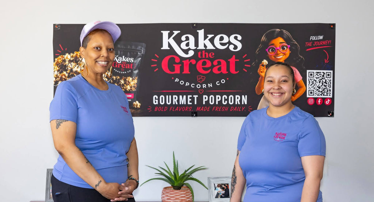 Alesha and Kaylynn standing in front of their Kakes the Great sign at a client event. 